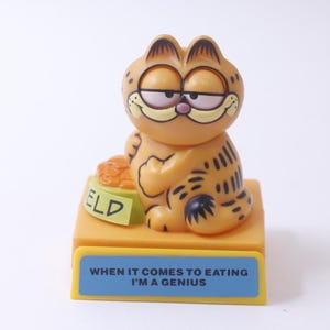May include: A Garfield figurine with an orange and black striped body. He is sitting on a yellow base with the words "When it comes to eating I'm a genius" on a blue background. The base also has a yellow bowl with the word "ELD" on it.
