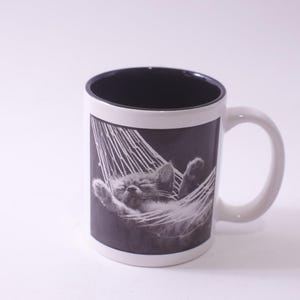 May include: White ceramic mug with a black interior and a white handle. The mug features a black and white image of a kitten sleeping in a hammock. The mug is a standard size for coffee or tea.