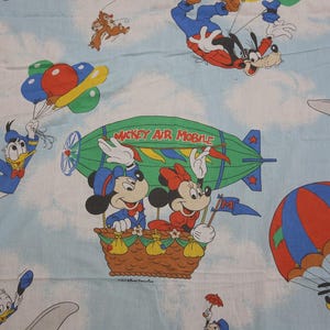 May include: Light blue fabric featuring a repeating pattern of Disney characters in the sky. Mickey and Minnie Mouse are in a hot air balloon, and Donald Duck is flying with a parachute. Other characters include Goofy and Pluto.