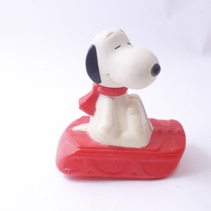 May include: A Snoopy figurine, featuring the iconic cartoon dog, is seated on a red sled. Snoopy is white with black ears and a black nose, wearing a red scarf. The sled is a bright red color.