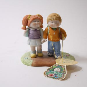 May include: A ceramic figurine of two Cabbage Patch Kids, a girl with orange pigtails and a boy with blond hair, standing together. The girl wears a purple top and white skirt, the boy an orange sweater and blue jeans. They stand on a small, grassy base.