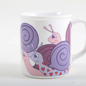May include: White ceramic mug featuring a whimsical design of several cartoon snails. The snails have purple and pink shells, with red heart details. The mug has a white handle and a simple, clean design.