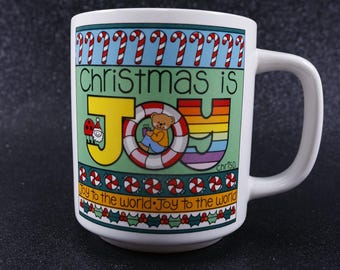 May include: White ceramic mug with a Christmas-themed design. The mug features the text "Christmas is JOY" in colorful letters, with candy canes, holly, and a teddy bear. The mug has a white handle.