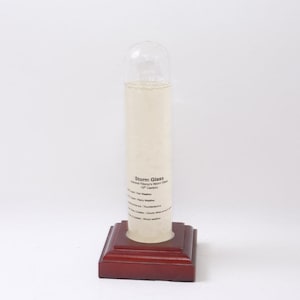 May include: A clear glass storm glass weather predictor with a cylindrical body and a rounded top. The glass tube is filled with a liquid and sits on a dark red wooden base. Text on the glass reads "Storm Glass".