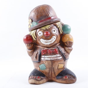 Op de afbeelding: Een keramieken clownfiguur met een bruine hoed en pak, met drie kleurrijke ballen. De clown heeft een rode neus, een rode strik en een lachend gezicht. Het beeldje is een decoratief item.