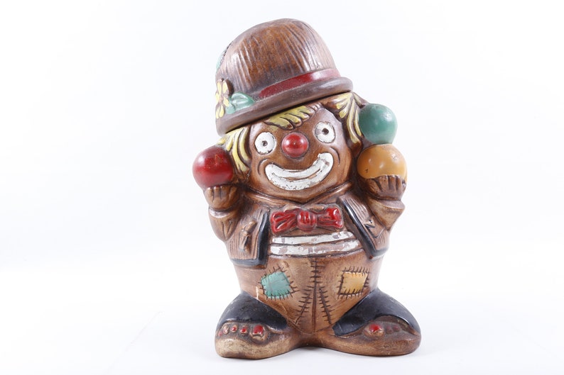 Op de afbeelding: Een keramieken clownfiguur met een bruine hoed en pak, met drie kleurrijke ballen. De clown heeft een rode neus, een rode strik en een lachend gezicht. Het beeldje is een decoratief item.