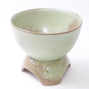 Pode incluir: Uma tigela de cerâmica verde claro com uma borda marrom fica em uma base de pedestal combinando com um design floral. A tigela é perfeita para servir lanches ou saladas.