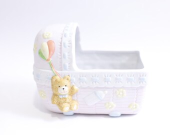May include: A white ceramic baby crib with a pink and blue trim. The crib has a teddy bear holding a balloon and a baby bottle on the side.