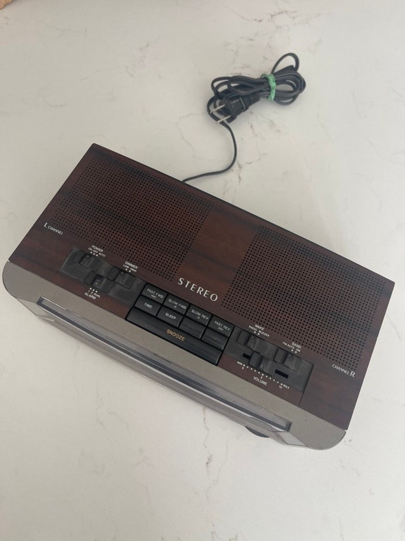 Pu&ograve; includere: Un radio sveglia stereo in legno marrone con un quadrante argentato e pulsanti neri. La radio ha un display digitale dell'orologio, un timer di spegnimento e un pulsante snooze. La radio ha anche un selettore di canali, una manopola del volume e un pulsante di accensione.