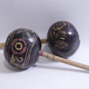 Pode incluir: Duas maracas de madeira com cabaças pintadas de preto e desenhos florais em dourado, rosa e verde. As maracas são conectadas por um pau de madeira.