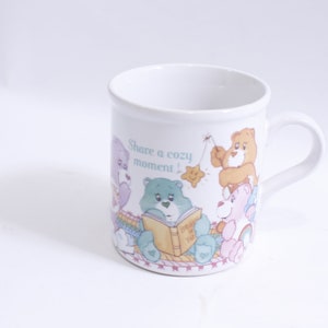 May include: White ceramic mug with a colorful illustration of Care Bears sharing a cozy moment. The mug features the text "Share a cozy moment!" and a Care Bear holding a book that says "Don't Worry, Be Happy!"