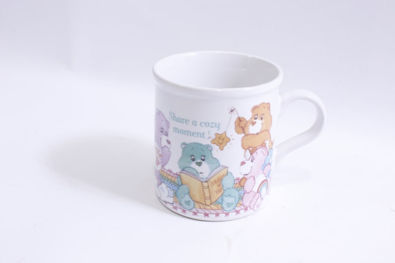May include: White ceramic mug with a colorful illustration of Care Bears sharing a cozy moment. The mug features the text "Share a cozy moment!" and a Care Bear holding a book that says "Don't Worry, Be Happy!"