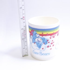 May include: White ceramic mug with a rainbow and hearts design featuring a blue Care Bear with a star on its chest. The mug says "Care Bears" at the bottom.