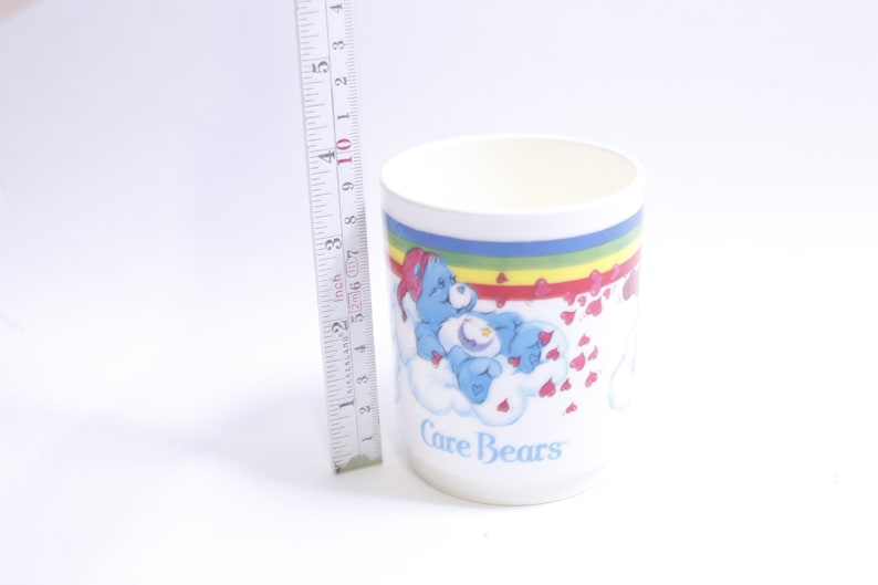 May include: White ceramic mug with a rainbow and hearts design featuring a blue Care Bear with a star on its chest. The mug says "Care Bears" at the bottom.
