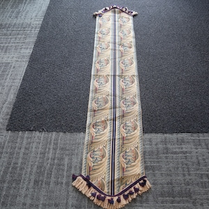 May include: A long, narrow table runner with a beige background and a repeating pattern of gold and purple leaves. The runner has a fringe border with purple tassels.