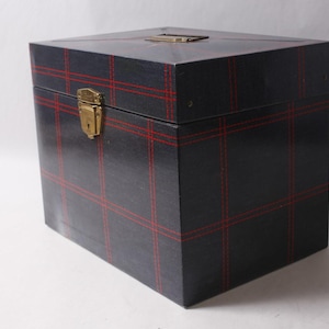 May include: A square storage box with a dark navy blue finish and a red grid pattern. The box features a gold-toned metal latch on the front and a matching gold-toned handle on the top lid. The construction is rigid with clean, sharp edges.