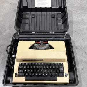 Könnte beinhalten: Eine Vintage-Schreibmaschine in Beige mit schwarzer Tastatur und Tasten, in einem schwarzen Tragekoffer. Die Schreibmaschine hat einen silbernen Wagen und rote Akzente. Der Koffer ist geöffnet und gibt die Komponenten der Maschine frei.
