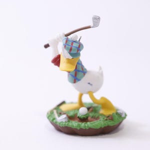 May include: A figurine of a white duck wearing a plaid hat and vest, swinging a golf club. The duck has a yellow beak and feet, and is standing on a miniature golf course with a golf ball.