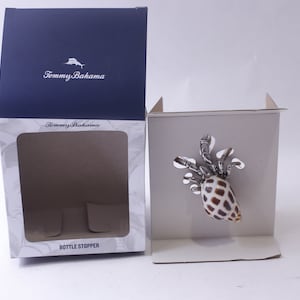 Könnte beinhalten: Ein Flaschenverschluss von Tommy Bahama in einer weißen Box mit einem transparenten Fenster. Der Verschluss zeigt eine silberne und braune Muschel mit einem silbernen Krabben-Design.