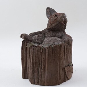 May include: A brown rabbit statue lounging in a textured, brown tree stump. The rabbit has its ears perked up and is resting its paws on the stump. The statue is a decorative item.