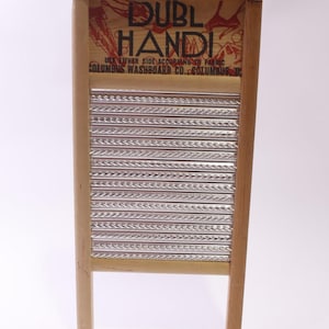 Op de afbeelding: Een vintage houten wasbord met een gegolfd metalen schrobvlak. Het bovenste gedeelte toont de tekst "DUBL HANDI" in zwart en rood. Het wasbord heeft een licht houten frame en is ontworpen voor het met de hand wassen van kleding.