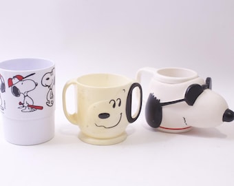 Può includere: Tre tazze a tema Snoopy. Una tazza bianca con grafica di baseball di Snoopy, una tazza giallo chiaro con la faccia di Snoopy e una tazza bianca a forma di Snoopy con occhiali da sole. Le tazze sono in ceramica.