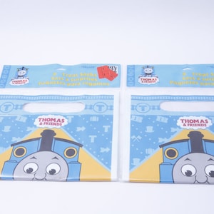 May include: Two blue and white Thomas & Friends treat bags with a handle. Each bag has a picture of Thomas the Tank Engine on the front. The bags are made of plastic and have a red and white striped design.