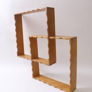 May include: A light brown, wooden wall shelf with a unique, interlocking design. The shelf features three rectangular sections, each with a wavy edge. The wood grain is visible, giving it a natural look. The shelf is designed to be mounted on a wall.