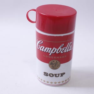 May include: Red and white Campbell's Soup thermos with a red lid and a handle. The thermos is shaped like a can of soup and has the Campbell's Soup logo and the words "Condensed Soup" on it.