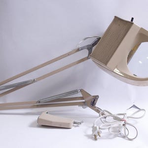 Op de afbeelding: Een beige vergrootlamp met een grote ronde lens en een verstelbare arm. De lamp heeft een rechthoekige kop met een geribbelde bovenkant en een klein handvat. De arm wordt ondersteund door veren en scharnieren. Een aparte beige voet en een wit netsnoer zijn ook zichtbaar.