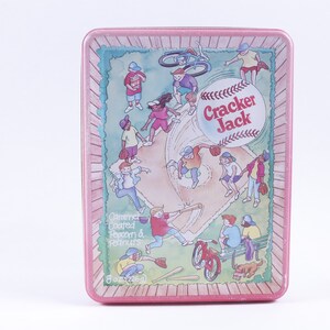 Puede incluir: Una lata rosa y verde con temática de béisbol. La lata presenta un diamante de béisbol con jugadores y una pelota de béisbol. El texto "Cracker Jack" está impreso en la lata. La lata es de 8 oz (226 g) y contiene palomitas de maíz y cacahuetes cubiertos de caramelo.