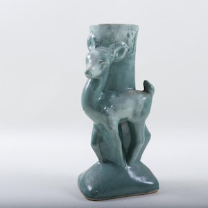 Könnte beinhalten: Eine teal-farbene Keramikvase mit einer Hirschfigur. Die Vase hat eine zylindrische Öffnung und einen Hirsch, der auf der Vorderseite modelliert ist. Der Hirsch steht auf einem abgerundeten Sockel. Die Vase ist etwa 20 cm hoch.