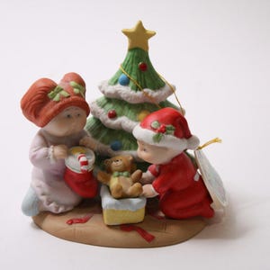 Peut inclure: Figurine de Noël en céramique avec deux personnages, un sapin de Noël et un ours en peluche. Un personnage porte un chapeau et un manteau rouges, l'autre une tenue rose et un chapeau rouge. L'arbre est vert avec des accents blancs et une étoile jaune.