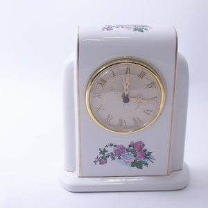 May include: White porcelain clock with a gold trim and Roman numeral clock face. The clock has a floral design with pink roses and green leaves.