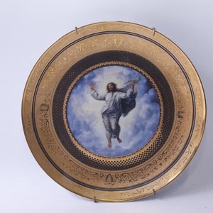 May include: A gold-rimmed porcelain plate with a blue and white image of a figure in a white robe ascending into the clouds. The plate has a black and gold decorative border.