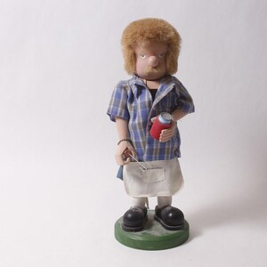 May include: A handcrafted figurine of a man with a red and blue plaid shirt, white apron, and black shoes. The figure has a red container in one hand and a tool in the other. The figurine stands on a green base.