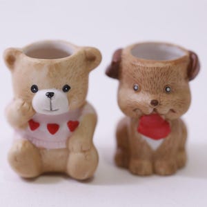 May include: Two ceramic figurines: a teddy bear and a dog. The bear is light brown with a pink sweater adorned with red hearts. The dog is brown with a red heart-shaped object. Both have open tops.