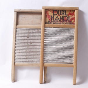 Op de afbeelding: Twee vintage houten wasborden. Eén wasbord heeft de tekst "DUBL HANDI" op een rode en crèmekleurige achtergrond. De wasborden hebben parallelle metalen schrobvlakken en zijn gemaakt van lichtgekleurd hout.
