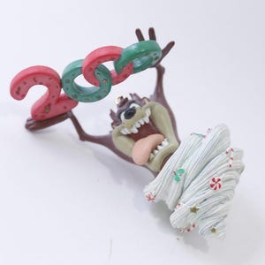 Könnte beinhalten: Ein Weihnachtsbaumschmuck mit einem Cartoon-Tasmanischen Teufel, der die Zahlen "2023" in Rot und Grün hochhält. Der Teufel trägt einen weißen, frostartigen Hut mit roten und weißen Zuckerstangen-Akzenten.