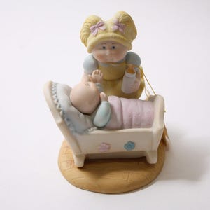 May include: A ceramic figurine depicting a doll-like figure holding a baby in a crib. The figure is wearing a yellow dress and holding a bottle. The baby is wrapped in a pink blanket. The base is a light brown color.
