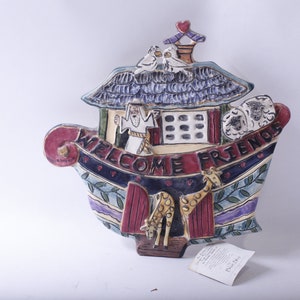Könnte beinhalten: Ein farbenfroher Wandbehang aus Keramik in Form eines Bootes mit den Worten "Welcome Friends" an der Seite. Das Boot hat ein Haus oben mit einem Schornstein und zwei Vögeln auf dem Dach. An der Seite des Bootes befinden sich zwei Giraffen und ein Schaf.