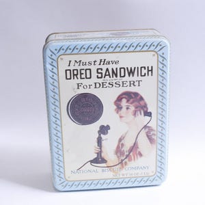 Op de afbeelding: Vintage rechthoekige blikken doos met een lichtblauwe rand en een crèmekleurig middenpaneel. Het paneel toont de tekst "I Must Have OREO SANDWICH For DESSERT" en een illustratie van een vrouw die een vintage telefoon vasthoudt. De blikken doos is gelabeld "NATIONAL BISCUIT COMPANY".