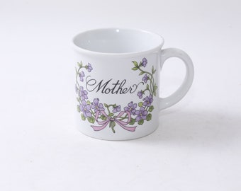 Könnte beinhalten: Weiße Keramiktasse mit Blumenmuster und dem Wort "Mutter" in schwarzer Schrift.