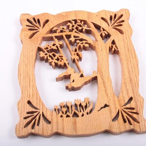 May include: Wooden decorative square with a carved tree and swing design. The light brown wood has a detailed cutout of a tree with a swing and floral accents. The edges have decorative cutouts.