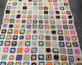 Puede incluir: Una manta de ganchillo colorida hecha de cuadrados de abuela en varios tonos de rosa, azul, verde, amarillo, naranja y rojo. Los cuadrados están unidos con hilo blanco, creando un efecto de patchwork.