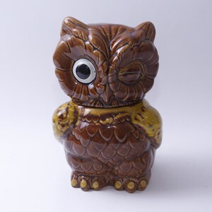 May include: A brown ceramic owl cookie jar with one eye open and the other eye closed, as if winking. The owl has yellow accents on its wings and feet.