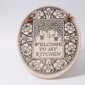May include: Oval ceramic kitchen sign with the text "WELCOME TO MY KITCHEN". The sign has a floral design with pink and green accents. It includes a brown rope for hanging.