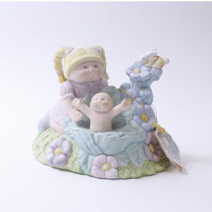 Puede incluir: Una figura de porcelana que representa a una niña con un vestido rosa y un bebé. La niña tiene coletas rubias y está sentada junto al bebé. La figura está decorada con colores pastel y flores, con un pequeño conejo encima.