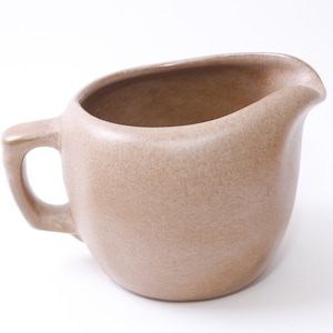 May include: A light brown ceramic creamer with a rounded body and a handle. The creamer has a speckled glaze.