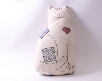 May include: A cream-colored cat-shaped pillow with embroidered details. The pillow features a red heart, patches with blue and floral patterns, and black outlines. The cat has a smiling face and is sitting upright.